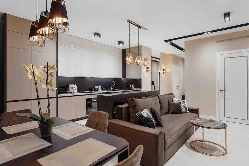 A modern living area features a comfortable sofa with decorative pillows, positioned beside a sleek dining table set for four. Warm lighting is provided by hanging fixtures, and a small table with soft accents is situated nearby, complementing the contemporary kitchen in the background.