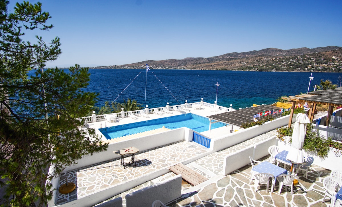 A panoramic view of a serene coastal landscape showcases a spacious salt-water pool surrounded by sun loungers. The white stone terrace features several outdoor seating areas, with the vibrant blue of the sea stretching towards the horizon under a clear sky.