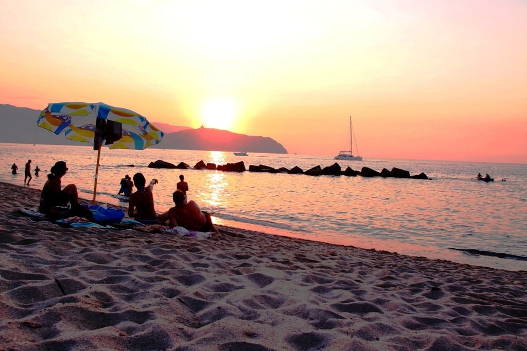 A sandy beach extends along the shoreline, where several individuals are seated under an umbrella. The sun sets in the background, casting a warm glow over the water. Silhouetted against the colorful sky, rocky formations and a distant sailboat are also visible.