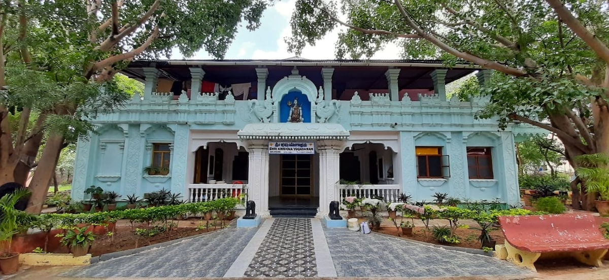 The front facade of Shri Krishna Yogashram is highlighted by intricate architectural detailing in pastel blue. Lush greenery surrounds the building, with a patterned pathway leading to the entrance. Statues and flowering plants are positioned on either side, adding to the serene atmosphere.