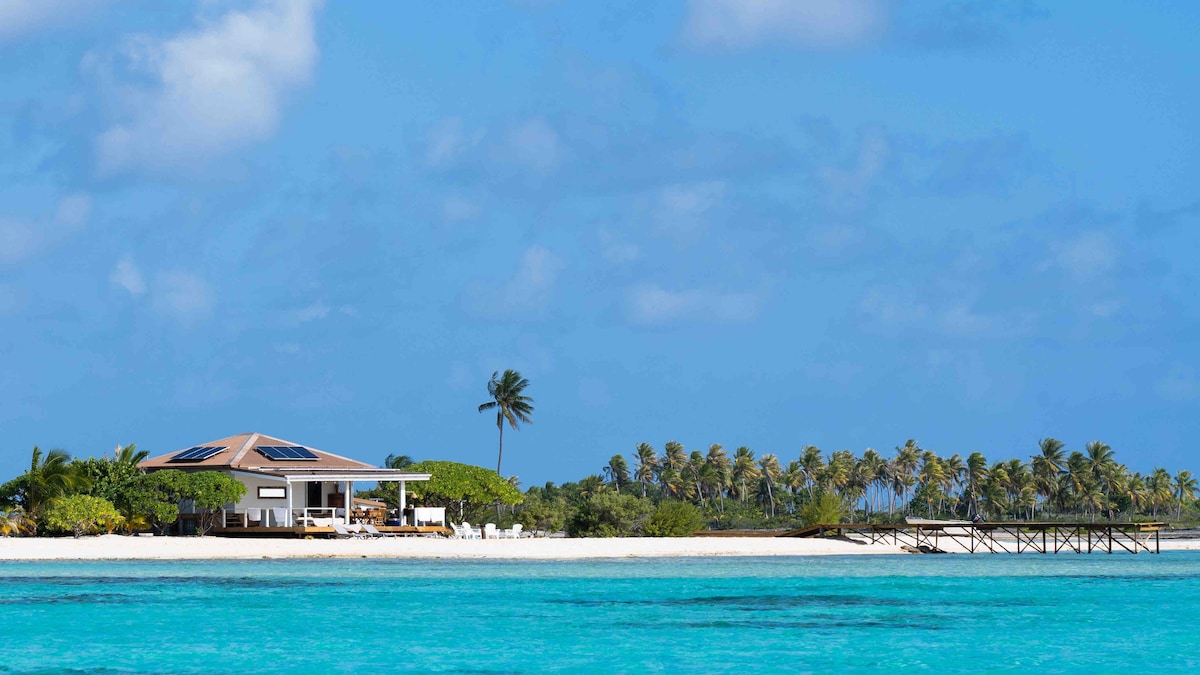 A comfortable eco-friendly house is situated on a private motu, surrounded by vibrant greenery and palm trees. The structure features a veranda overlooking a turquoise lagoon, with a wooden dock extending into the water, enhancing the tranquil island atmosphere.