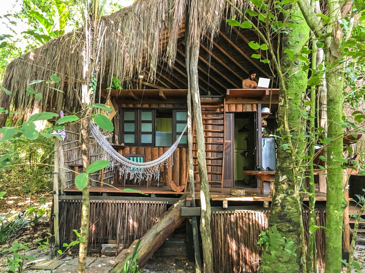 A wooden bungalow with a thatched roof is surrounded by lush greenery. A hammock hangs on the porch, where seating options are also visible. There are stairs leading up to the entrance, and the natural materials blend harmoniously with the surrounding environment.
