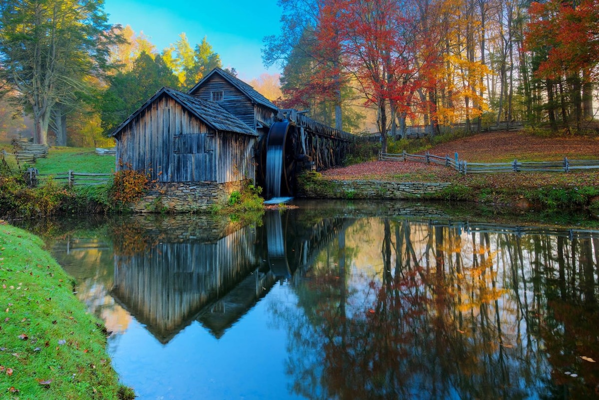 A historic wooden mill with a water wheel rests beside a tranquil pond, reflecting the vibrant autumn foliage and surrounding trees. The landscape is accented by a lush green lawn, creating a serene atmosphere typical of the Blue Ridge Mountains.