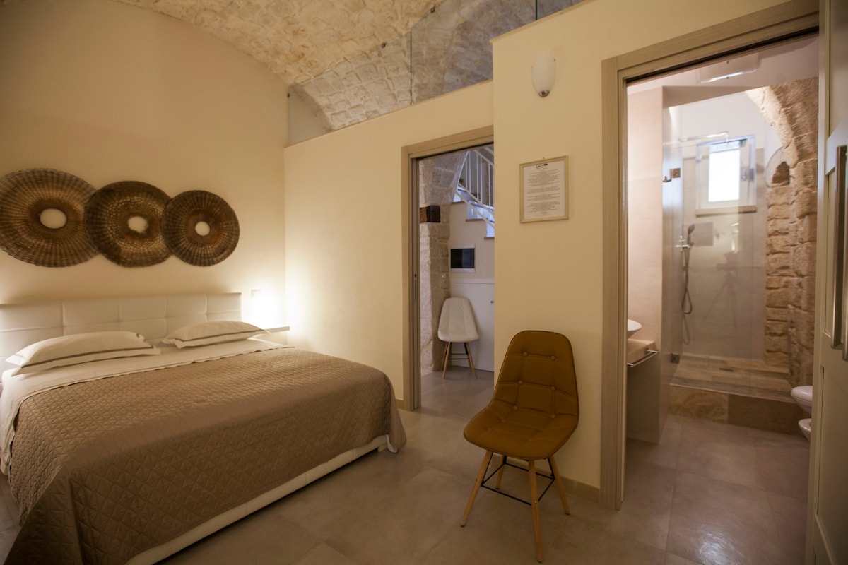 A room features a spacious bed with a neutral-colored quilt and two bedside lights. On the wall, three circular decorative elements are displayed. A bathroom is visible through an open door, showcasing a tiled shower and natural stone accents.