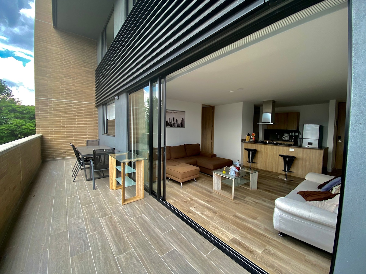 A spacious terrace is visible, featuring a dining table and chairs, along with a comfortable seating area. Inside, an open-plan layout showcases a well-equipped kitchen, a large sofa, and bar stools. Large glass doors enhance the connection between indoor and outdoor spaces.