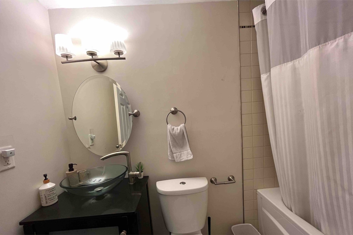 A modern bathroom features a glass vessel sink complemented by a sleek black countertop. A round mirror is mounted above the sink, and a white towel hangs nearby. The space includes a white tub and a shower curtain, creating a clean and functional design.
