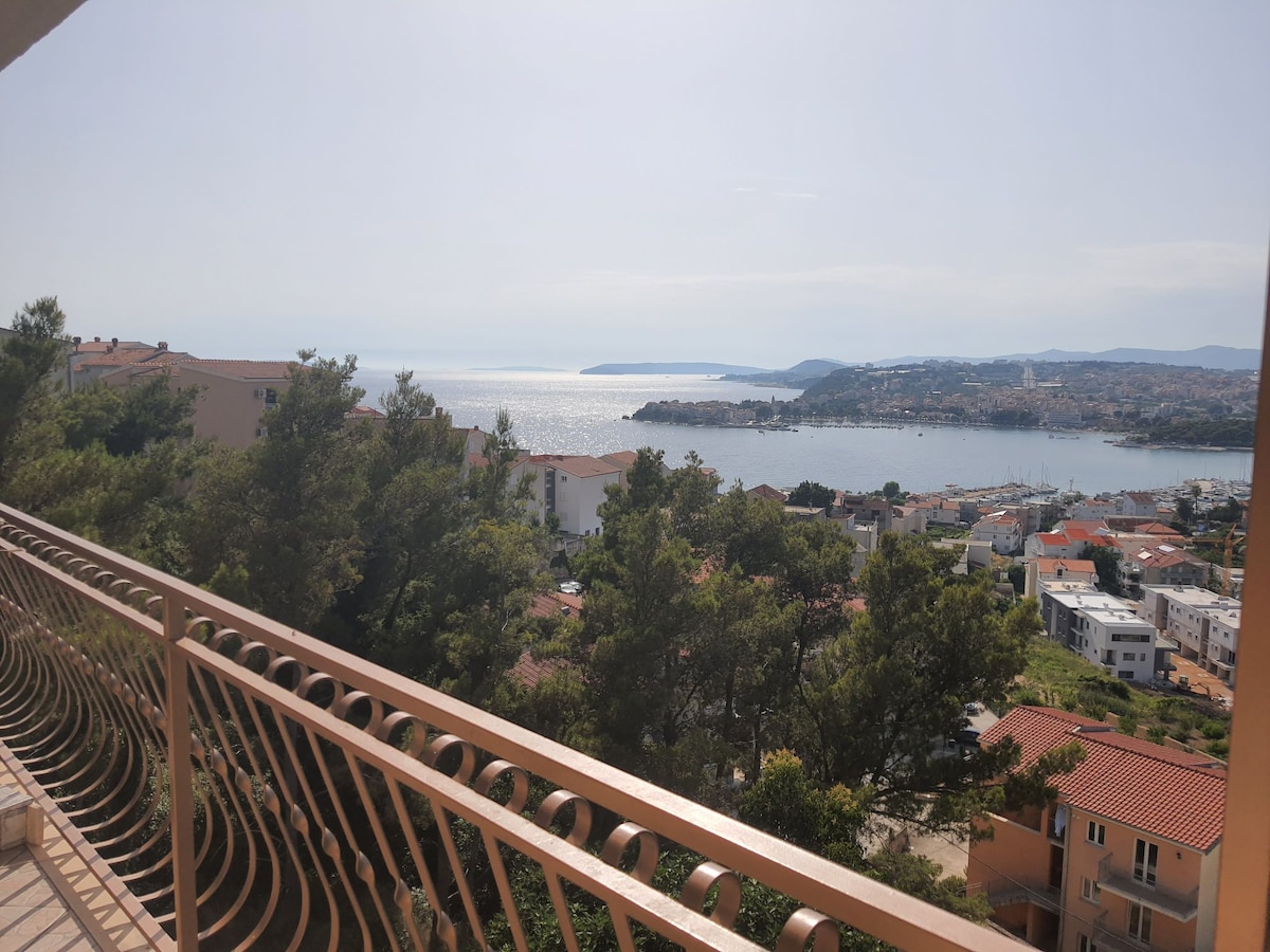 A scenic view of the coastline is displayed, featuring a sparkling sea and distant islands. The image captures a blend of lush greenery and terracotta-roofed buildings nestled along the hillside, with a balcony railing framing the expansive panorama.