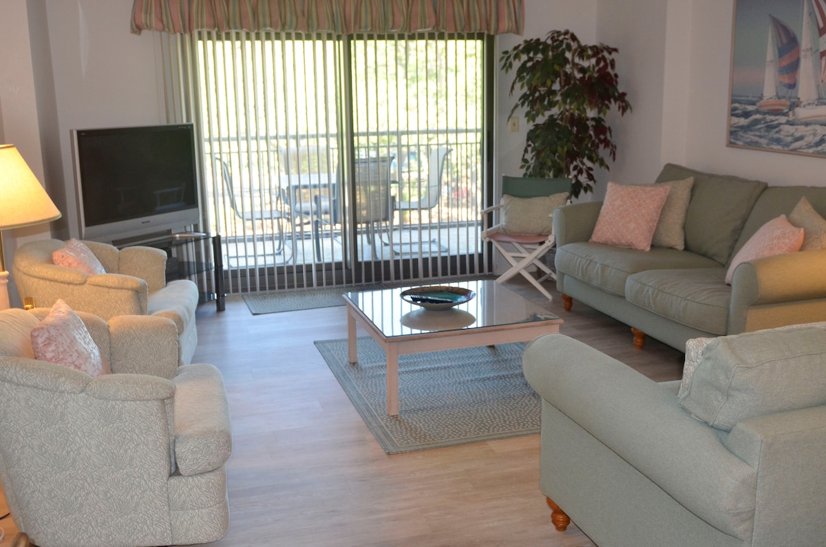 A comfortable living area features two light-colored sofas and two armchairs arranged around a coffee table. Large sliding glass doors allow natural light and provide a view of the outdoor seating area, with vertical blinds partially drawn. Soft pastel accents contribute to a relaxed ambiance.