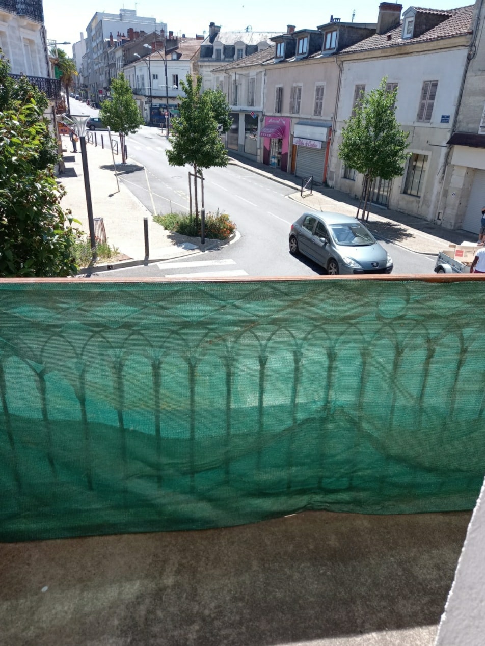 A view from a balcony overlooks a quiet street lined with small trees and buildings. A green protective netting is visible in the foreground, partially obscuring the outdoor space. A silver car is parked along the roadside in the distance.