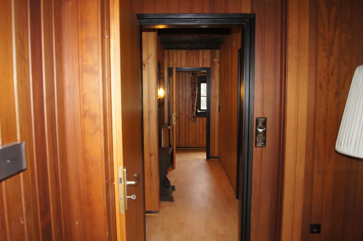 A hallway with wooden paneling features doors leading to various rooms. Soft lighting illuminates the space, highlighting the warmth of the wood. The floor is finished in a light tone, providing contrast to the darker walls. A visible light switch allows for easy access to lighting.