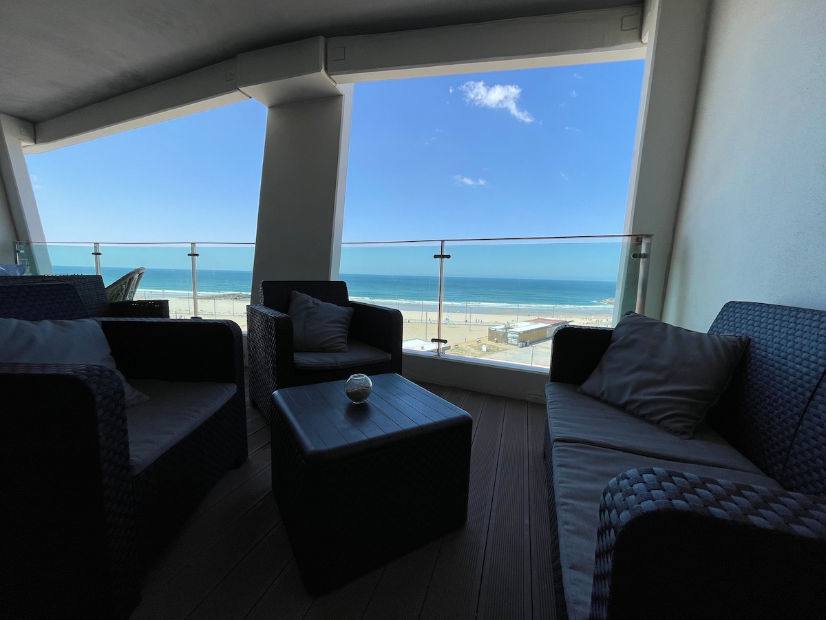 A modern balcony is displayed with comfortable wicker seating and a small table. The sea view is visible through large glass panels, showcasing the horizon and beach. Natural light brightens the space, creating an inviting spot for relaxation.