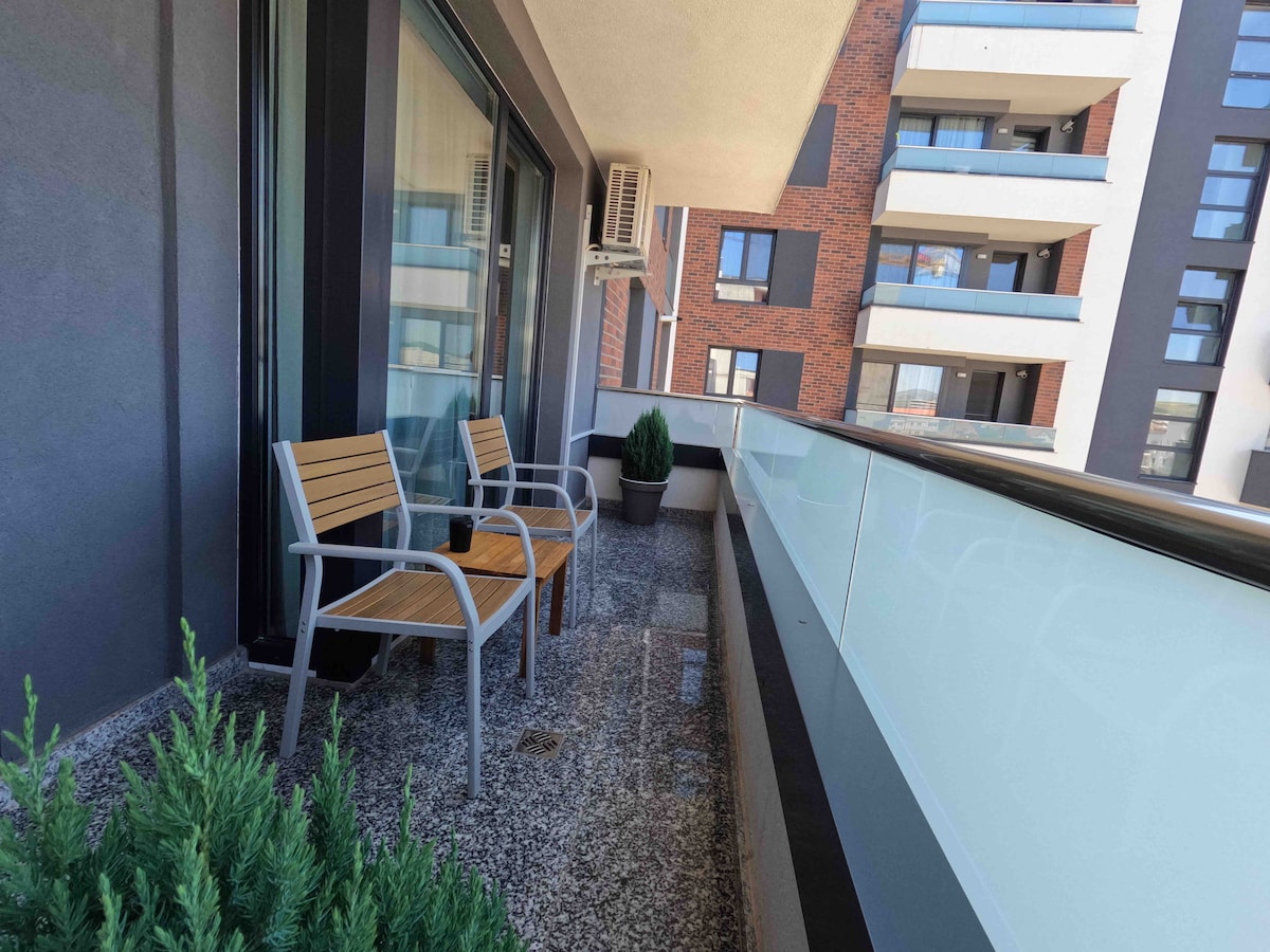 A generous terrace is furnished with two wooden chairs, providing a cozy spot to enjoy the outdoors. The space is surrounded by greenery and features a glass railing, enhancing the views of the surrounding architecture.