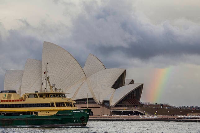 A green ferry glides through the water near the iconic Sydney Opera House, characterized by its unique shell-like architecture. In the background, a faint rainbow arcs across a cloudy sky, adding a subtle splash of color to the serene harbor scene.