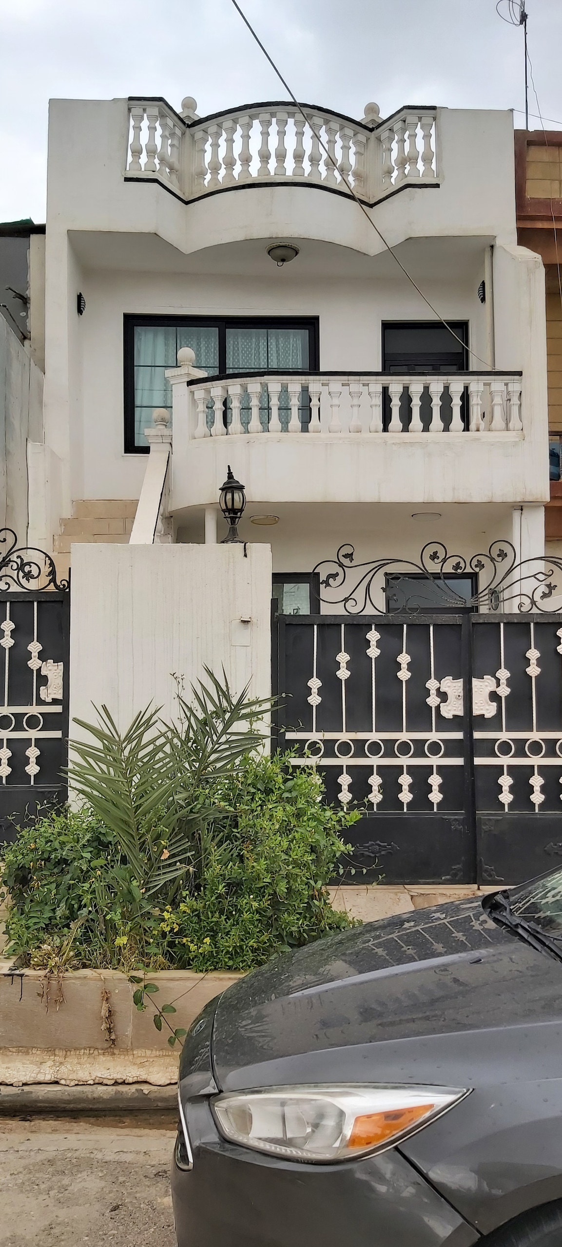 A modern two-story house is shown, featuring a balcony with decorative railing above the entrance. A neatly landscaped area with greenery is situated in front of the black gate. A parked gray car is visible in the foreground.