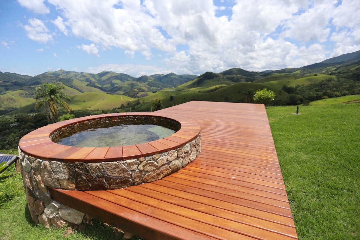 A circular hot tub is surrounded by a wooden deck, offering views of rolling green hills and open skies. The structure is crafted with a mix of smooth wood and natural stone, blending with the surrounding landscape.