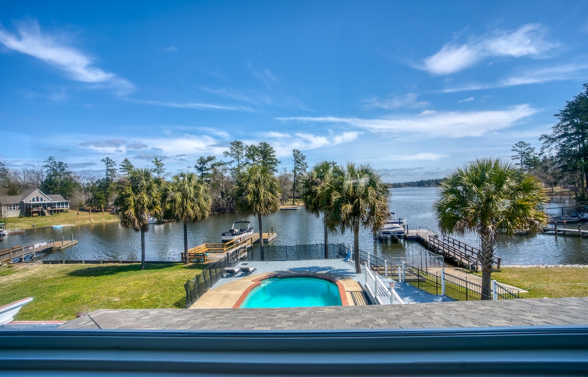 A scenic view of the lake from the property includes palm trees lining the shore and inviting blue water, with a pool visible in the foreground. Series of docks extend into the water, providing access for boating activities.