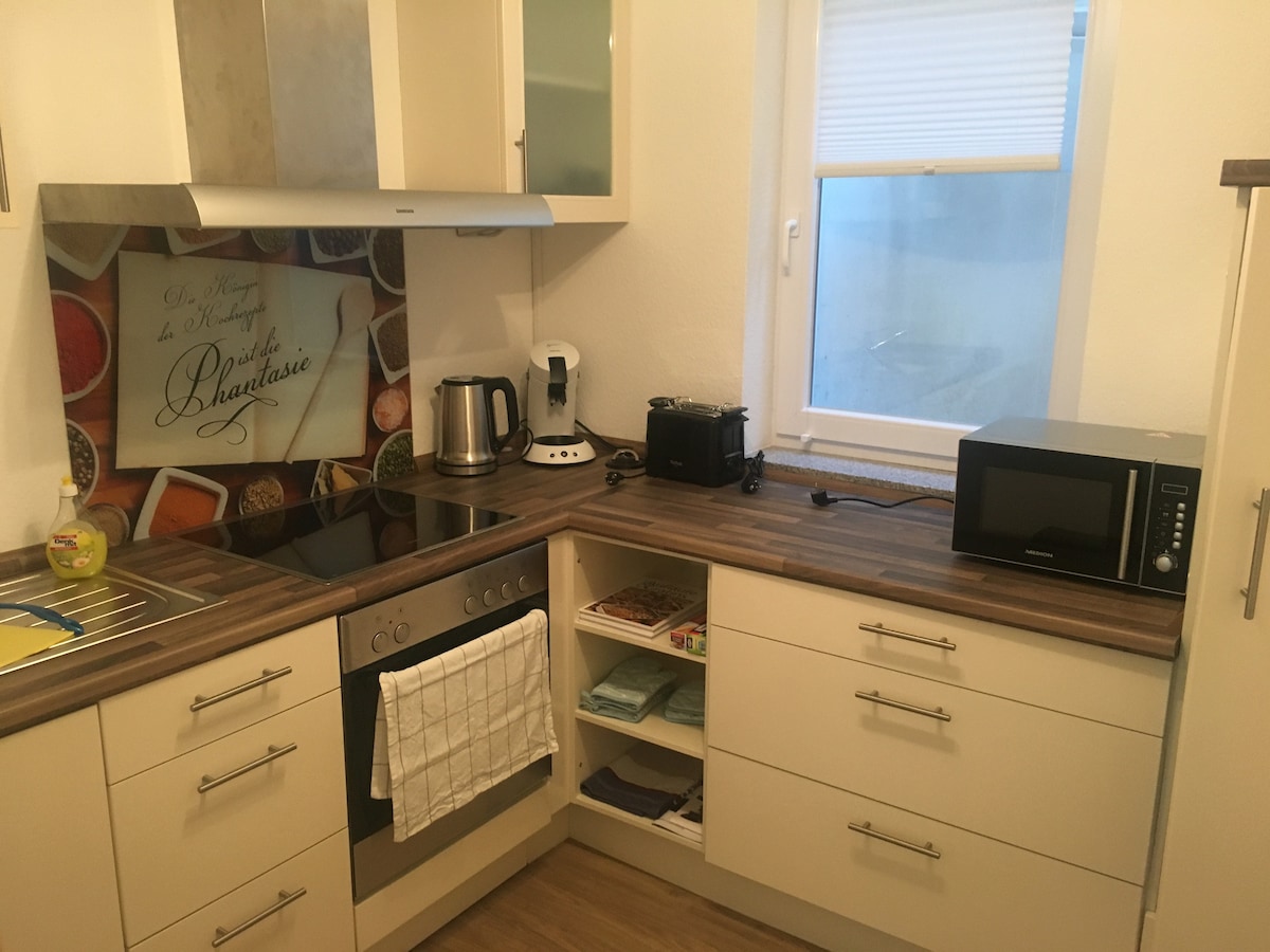 A well-equipped kitchen is visible, featuring modern cabinetry and a wooden countertop. Included appliances are a kettle, coffee machine, toaster, and microwave. A decorative backsplash with colorful food imagery adds interest. Natural light enters through a window, enhancing the functional layout.