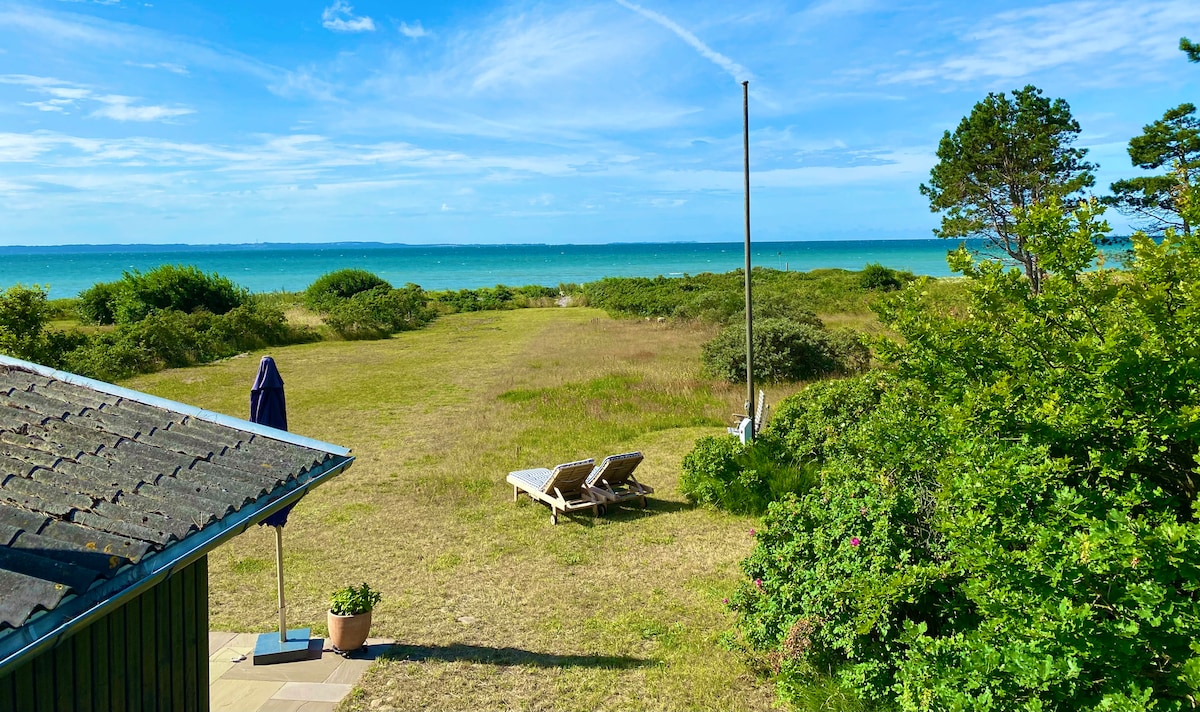 A serene outdoor view highlights a grassy area leading to the beach. Two lounge chairs are positioned for relaxation, with the calm turquoise sea and distant horizon visible. Lush green shrubs and trees frame the landscape under a bright blue sky.