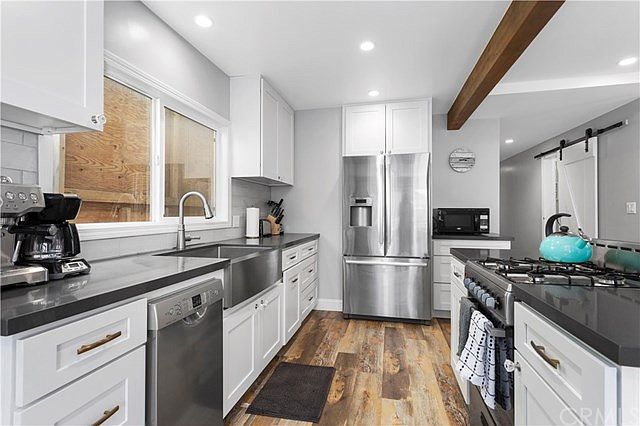 The kitchen features sleek white cabinetry and stainless steel appliances. A double sink is positioned under a window, and a modern gas stove is visible on one side. Countertops are spacious, and a coffee maker and microwave are included for convenience.