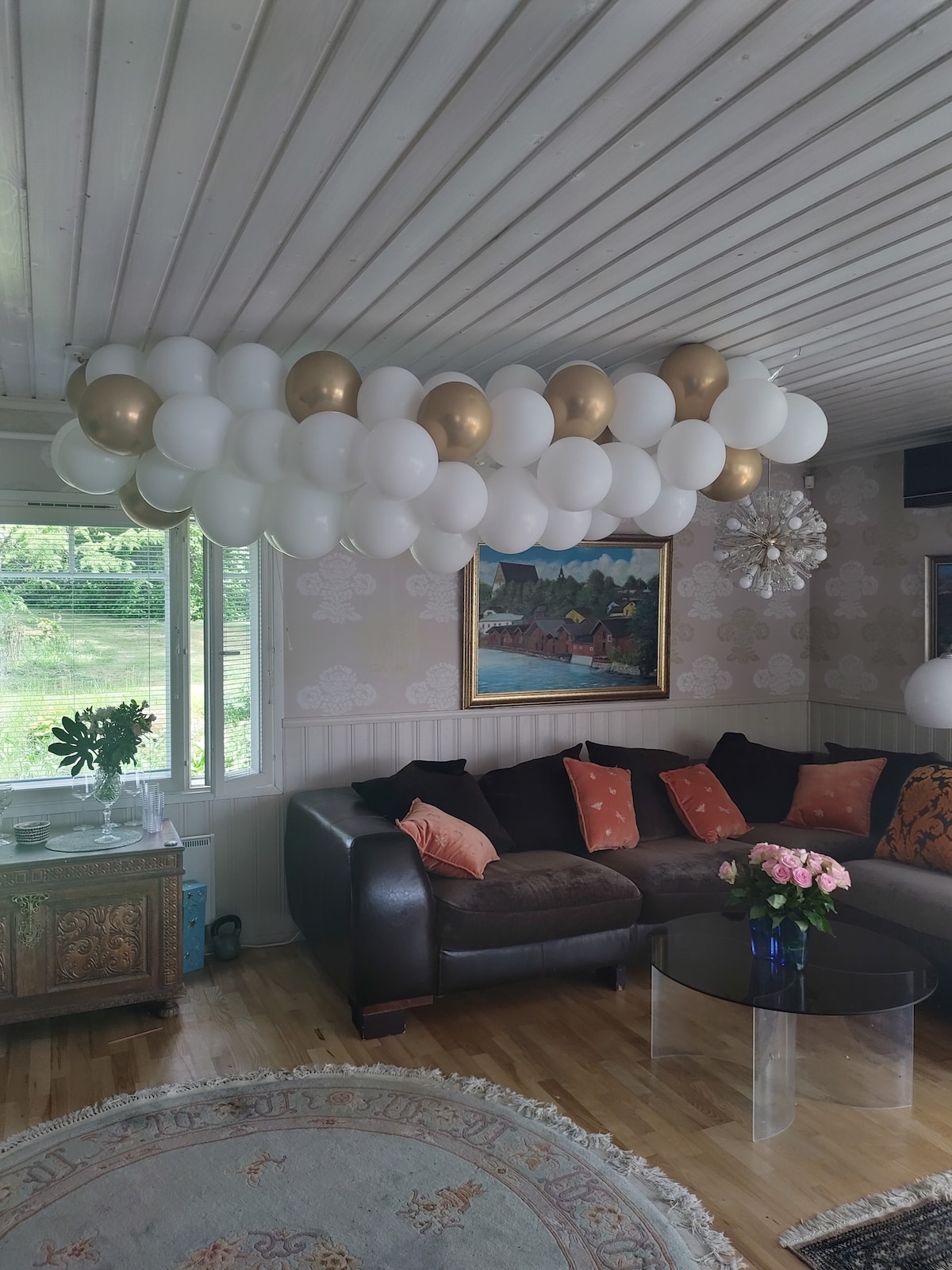 A living area is accented by a creative arrangement of white and gold balloons suspended from the ceiling. A large, dark leather sectional sofa features decorative pillows, inviting comfort. A round glass coffee table sits in front, and a colorful painting adorns the wall behind.