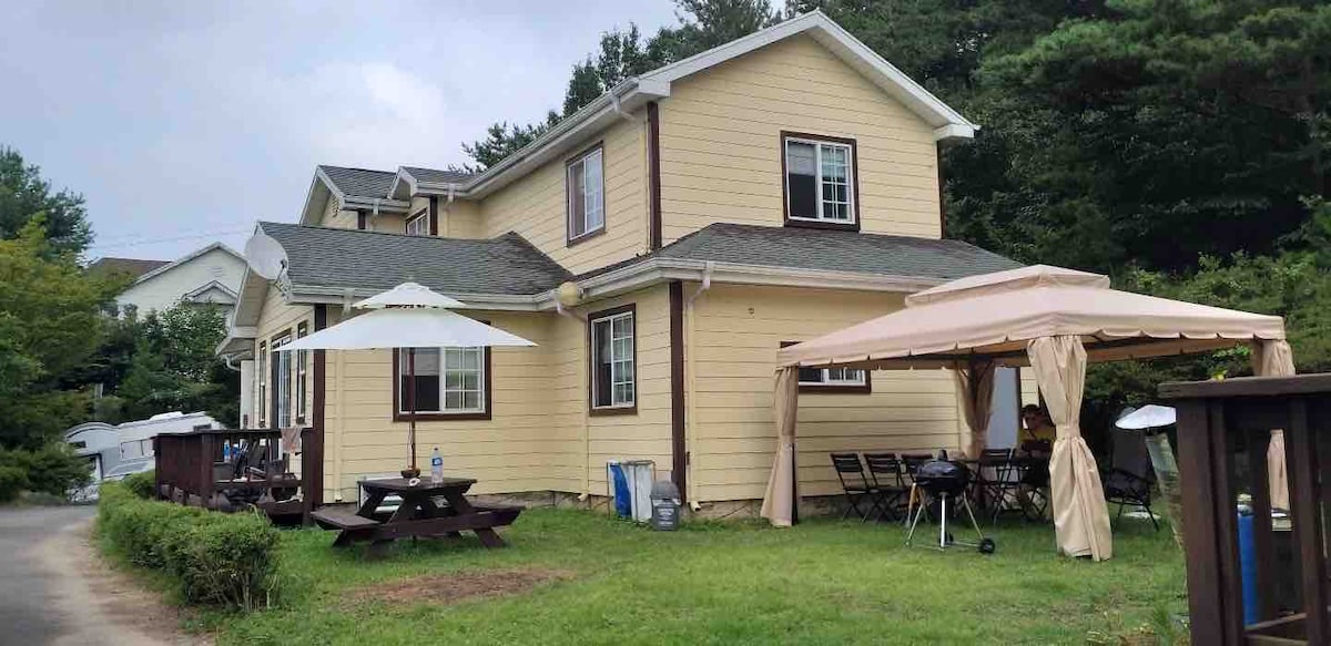 A charming wooden house with yellow siding is complemented by a spacious outdoor area. A covered shelter and a large umbrella provide shade for tables and chairs, while a barbecue grill is positioned nearby, all set against a backdrop of lush greenery.