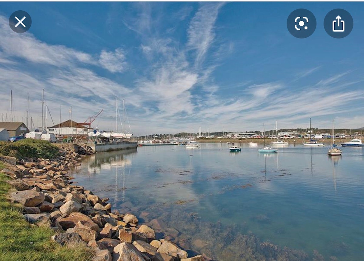 A serene waterfront scene displays calm waters reflecting the clear blue sky. Various boats are moored nearby, while a rocky shoreline is visible in the foreground. The background features buildings and masts, indicating an active marina environment.