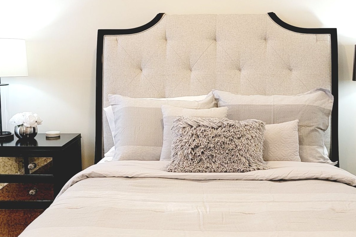 A spacious bedroom features a plush headboard with a tufted design, complemented by light-colored bedding. A small decorative pillow adds texture, while bedside lamps provide a soft glow. A mirrored nightstand stands beside the bed, reflecting light and enhancing the room's elegance.