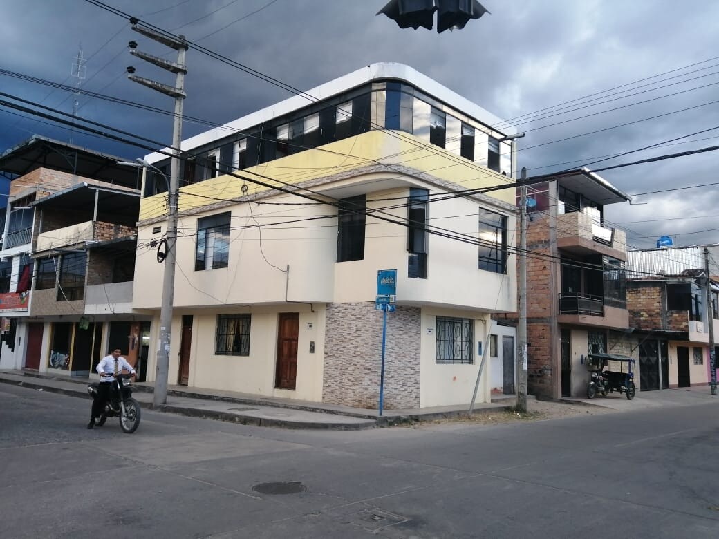 The exterior of a two-story house is shown, featuring a combination of light and dark beige walls. Large windows occupy the upper level, while the ground floor has a stone accent. Nearby streets and surrounding buildings are visible, along with overhead power lines.