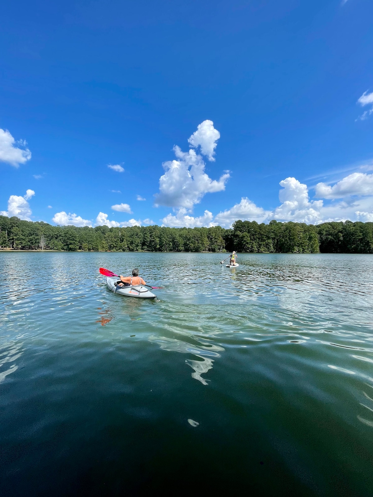 A serene lake setting features two individuals engaging in water activities. One person paddles a kayak with a red paddle while the other stands on a paddleboard. The clear blue sky is dotted with fluffy white clouds, and the lush greenery of the shoreline surrounds the calm water.