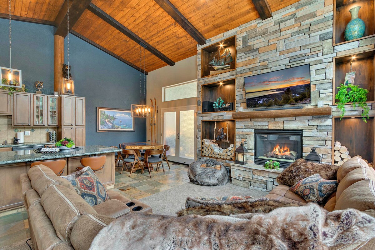 The spacious living area features a large sectional sofa, a stone gas fireplace, and a big screen TV mounted above a rustic stone wall. Natural light floods the space through ample windows, highlighting the wood-beamed ceiling and inviting atmosphere.