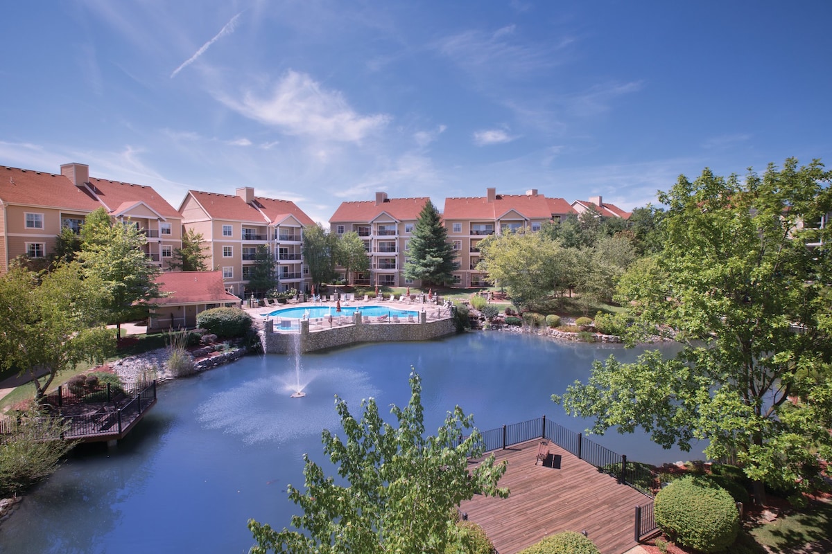 A scenic view of a resort property is captured, featuring a serene pond with a fountain and lush greenery. Surrounding buildings provide a backdrop, while a pool is visible nearby, inviting relaxation amidst the natural landscape.