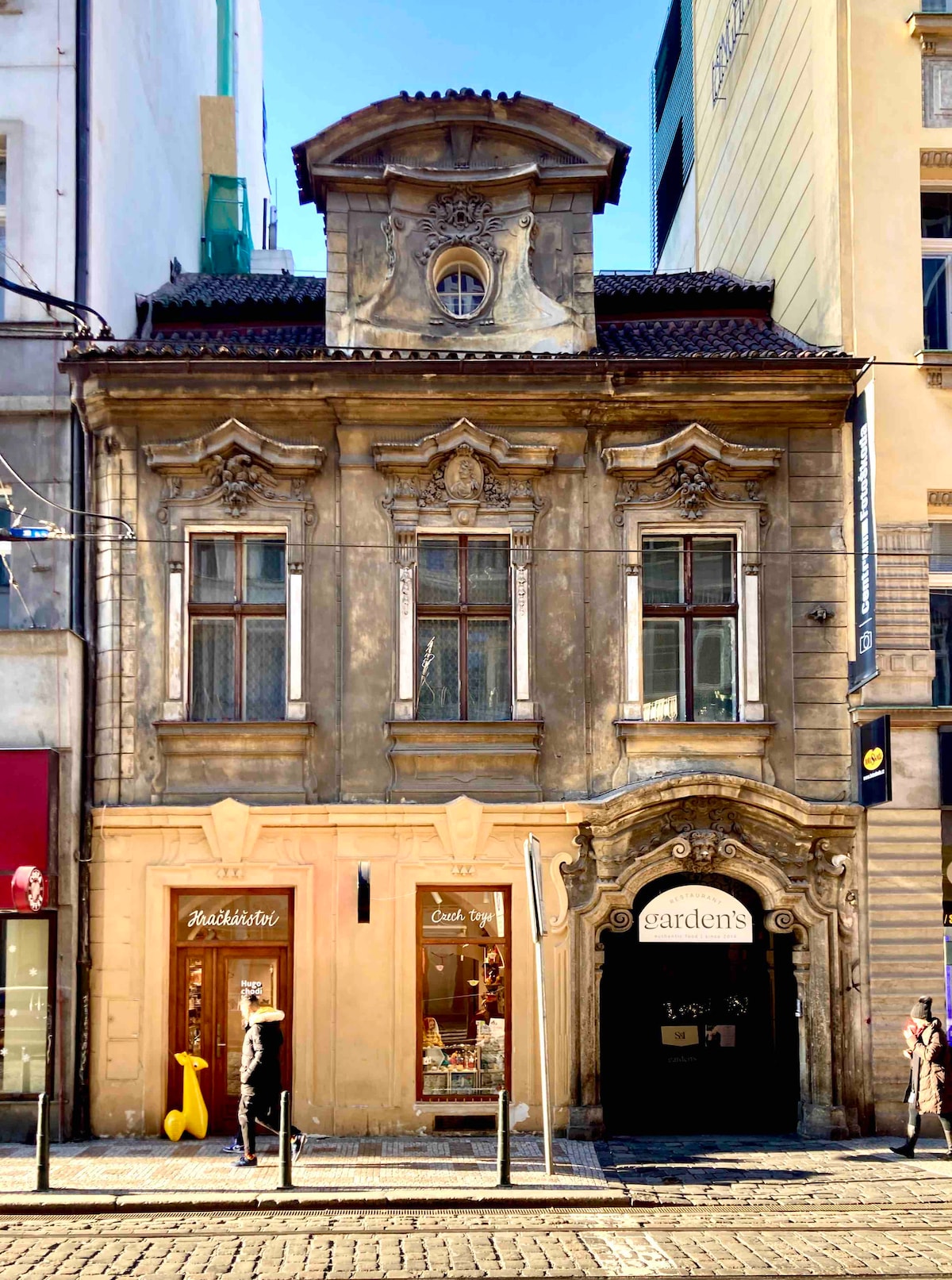 A historic building displays a unique architectural design with ornate details. Large windows are framed by decorative elements, and an elegant clock is positioned above the entrance. The facade is complemented by small shops on the ground floor, inviting exploration in the vibrant surrounding area.