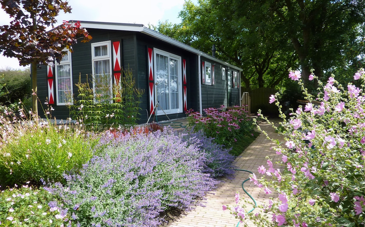 A charming chalet is surrounded by colorful flowering plants and greenery. Large glass doors provide a view into the interior, while a pathway lined with blooming flowers leads to the entrance. The building is adorned with decorative red accents on the exterior.