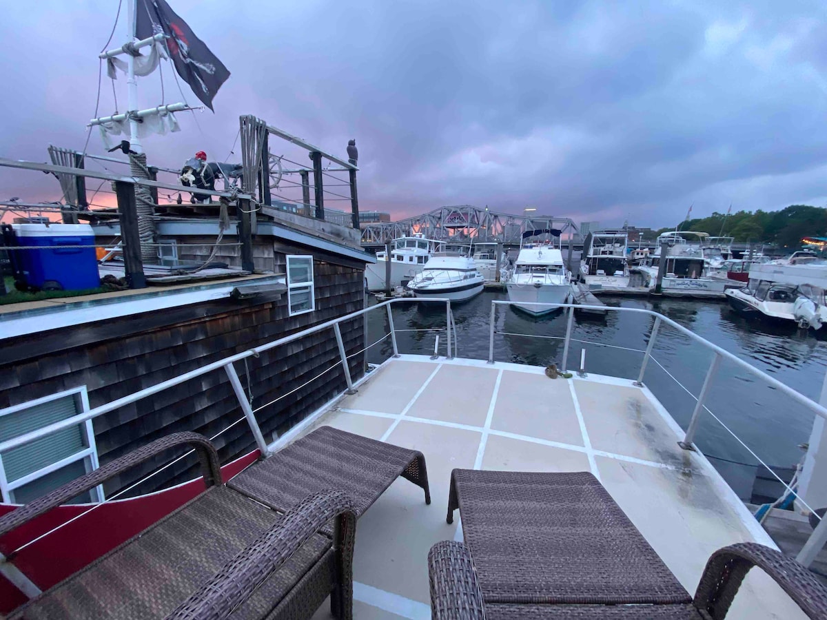 An upper deck is shown with two woven lounge chairs positioned for relaxation. The surrounding marina is visible, with several boats anchored nearby. A cloudy sky casts a serene atmosphere over the water, creating a tranquil setting.