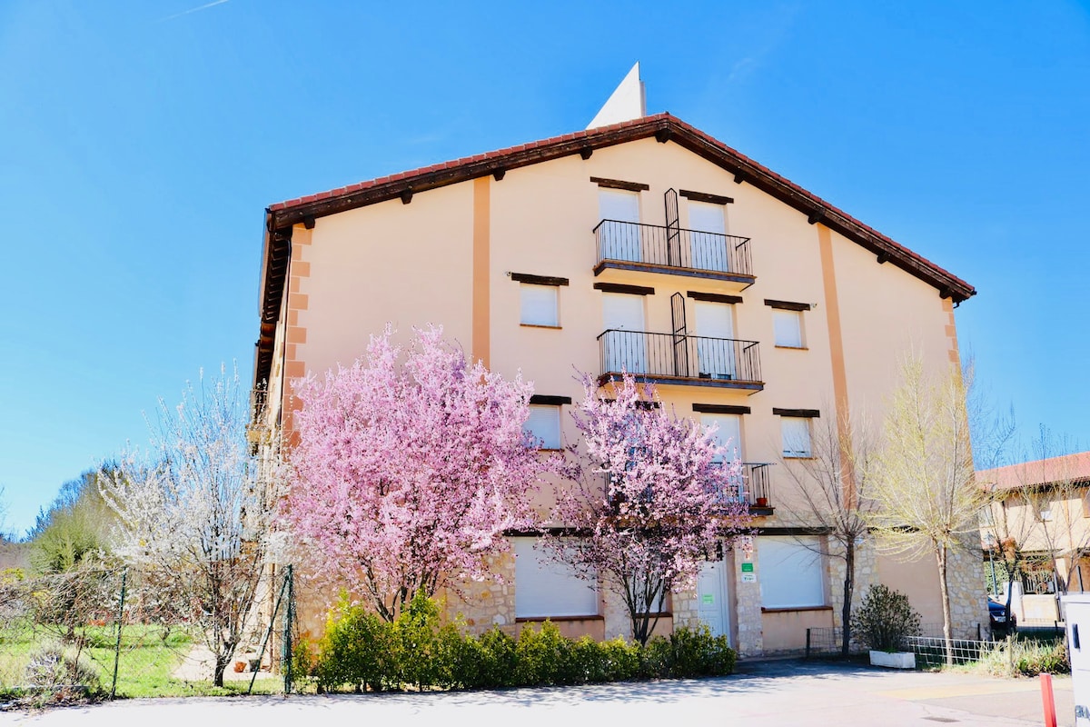 A rustic two-story building is framed by blooming trees, showcasing pink blossoms in the foreground. The structure features multiple balconies and a triangular roof, with well-maintained greenery surrounding the entrance, under a clear blue sky.