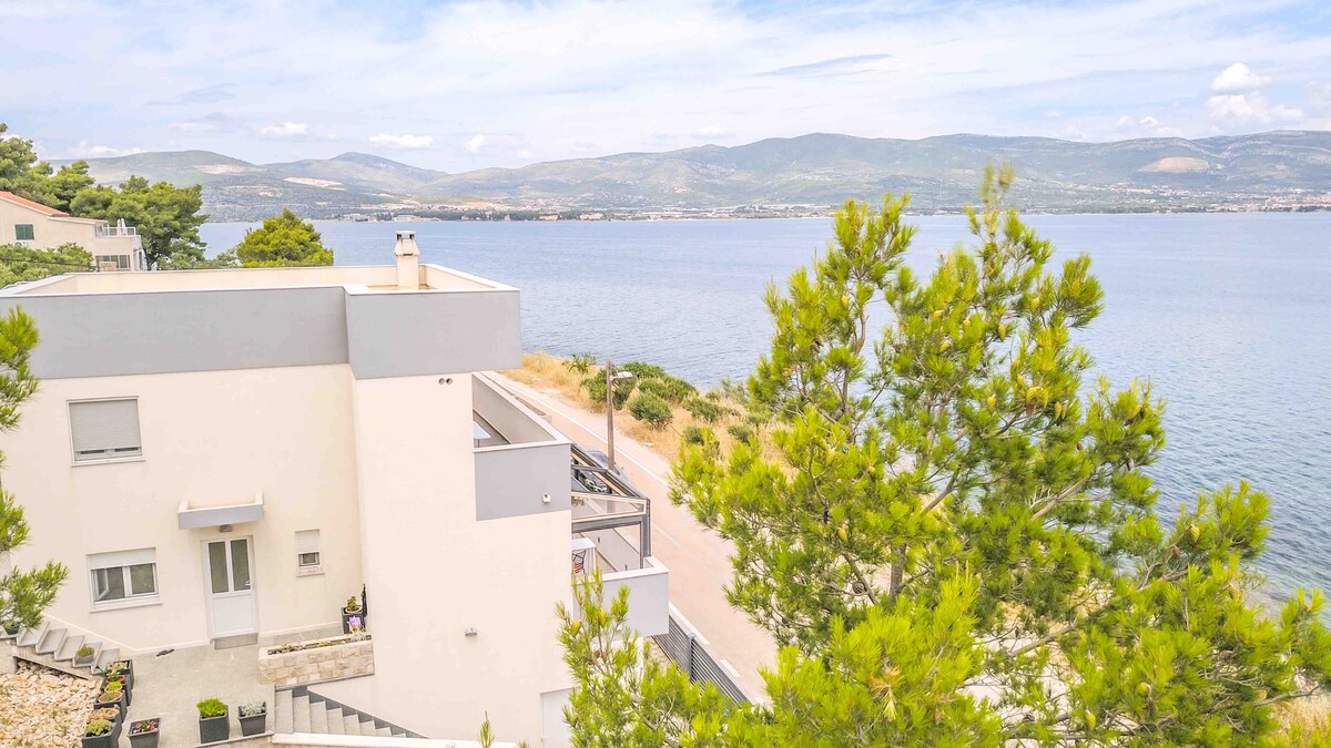 A modern apartment building is visible from an elevated angle, situated along the coastline. The blue sea is seen stretching towards the horizon, with distant mountains providing a scenic backdrop. Green trees frame the view, enhancing the natural setting.
