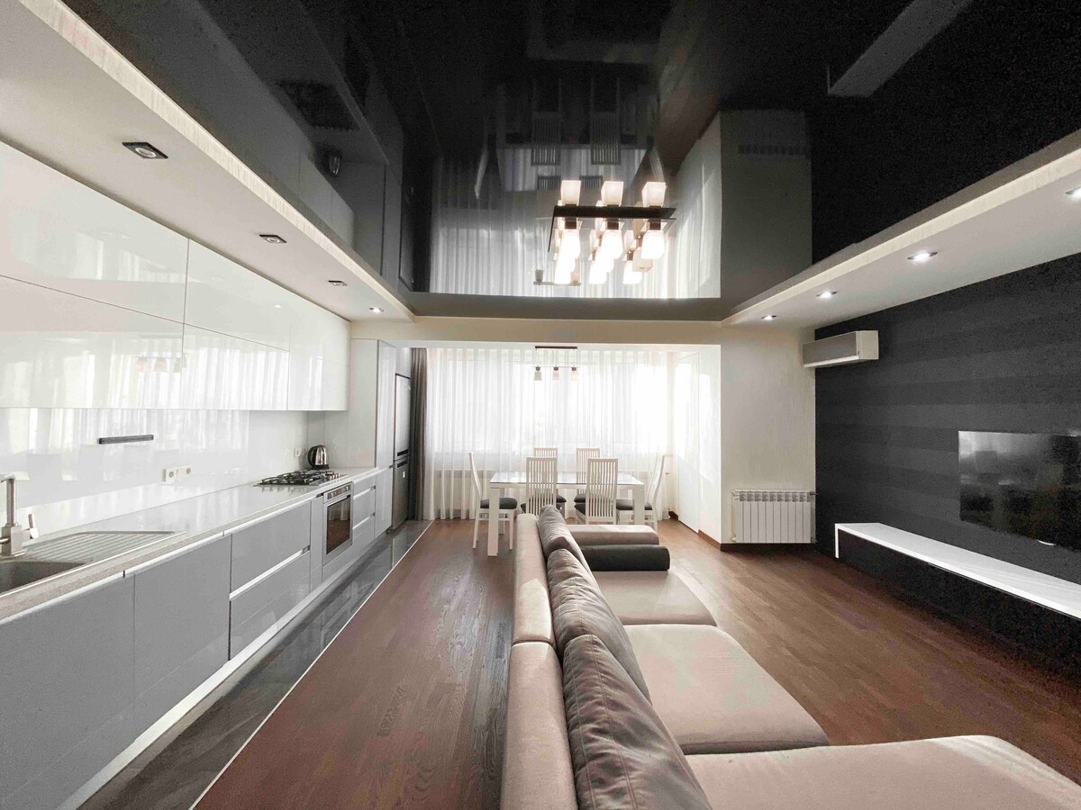 An open living space showcases modern white cabinetry and stainless-steel appliances in the kitchen area. A sectional sofa is positioned centrally on polished wood flooring. Natural light filters through sheer curtains, illuminating the dining area with a contemporary chandelier above.