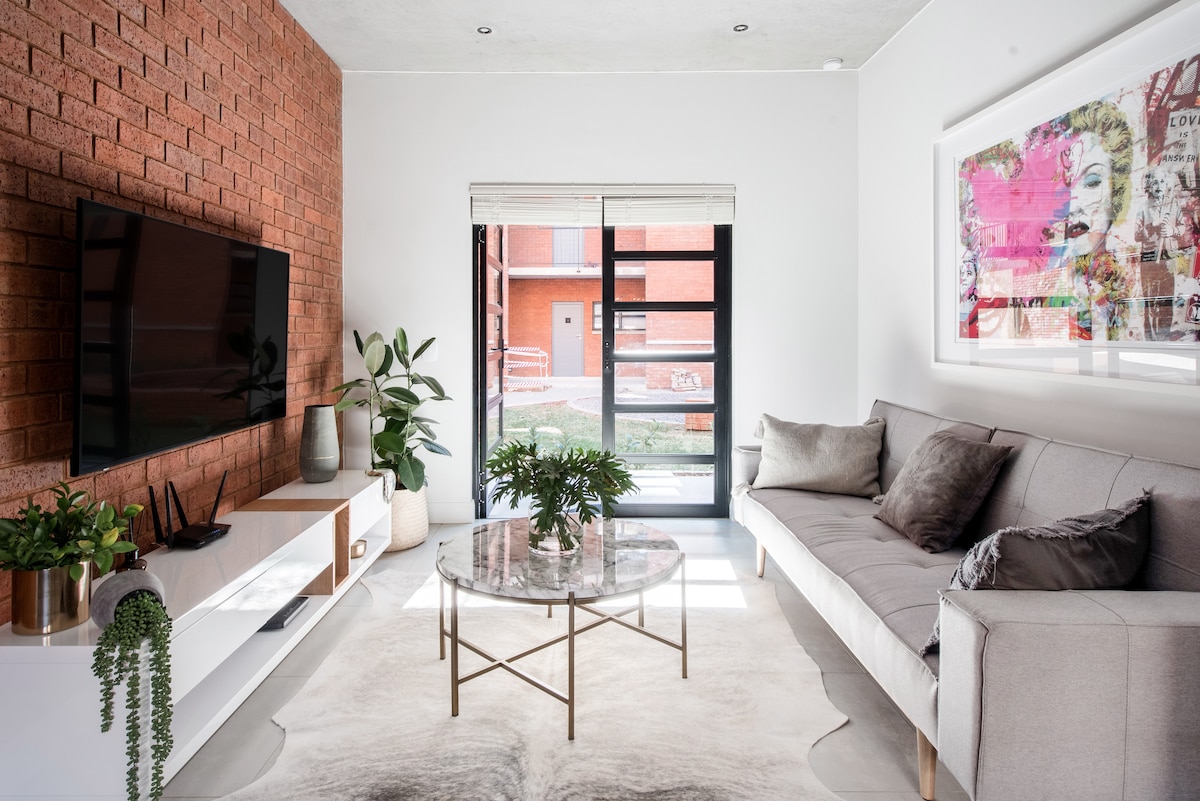 A modern living area features a light-colored sofa, a round coffee table with a marble top, and a flat-screen television mounted on a brick wall. Potted plants are placed around, and colorful artwork is displayed above the sofa, accentuating the space.