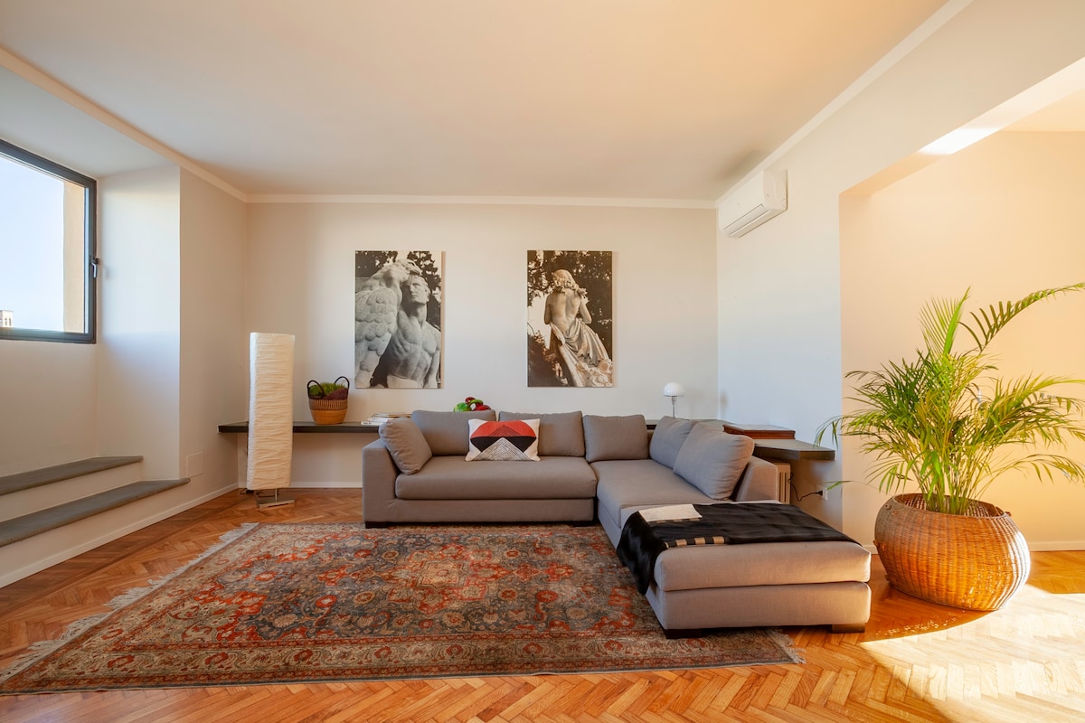A spacious living area features a comfortable gray sectional sofa accompanied by a plush throw. Two large artworks adorn the walls above, and a fern plant in a decorative pot adds a touch of greenery. Natural light floods through a large window, highlighting the wooden floor and minimalist decor.