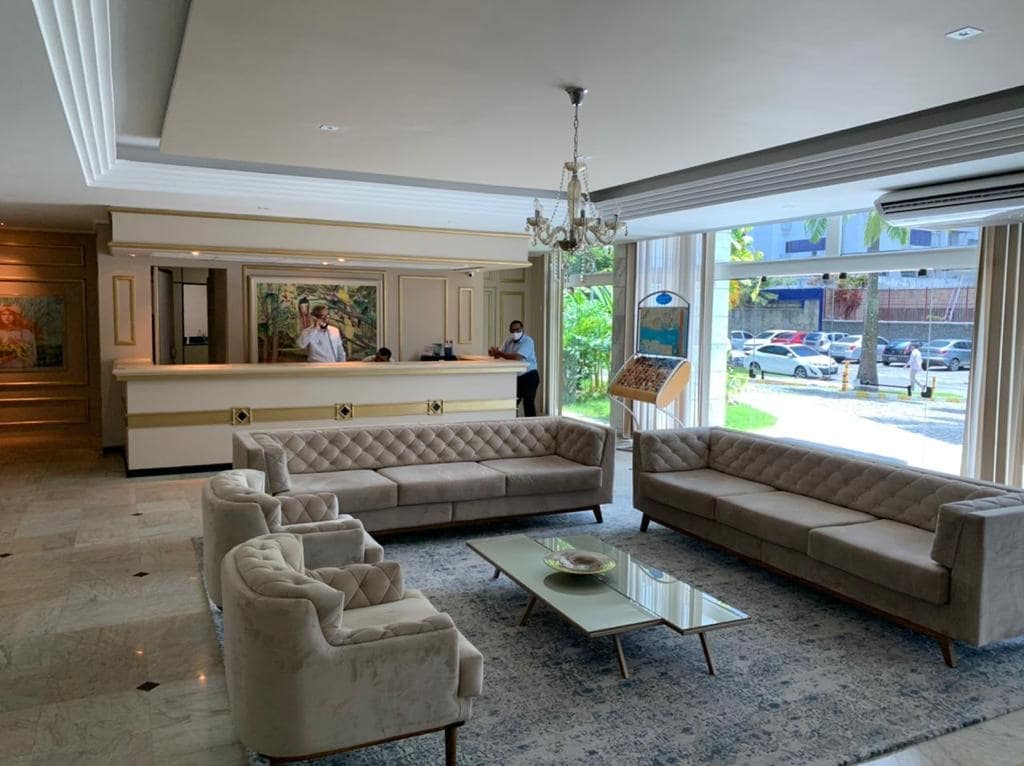 A spacious lobby is presented, featuring elegant tufted sofas arranged around a central coffee table. A reception area is visible in the background, attended by staff. Large windows allow natural light to fill the space, complementing the marble flooring.