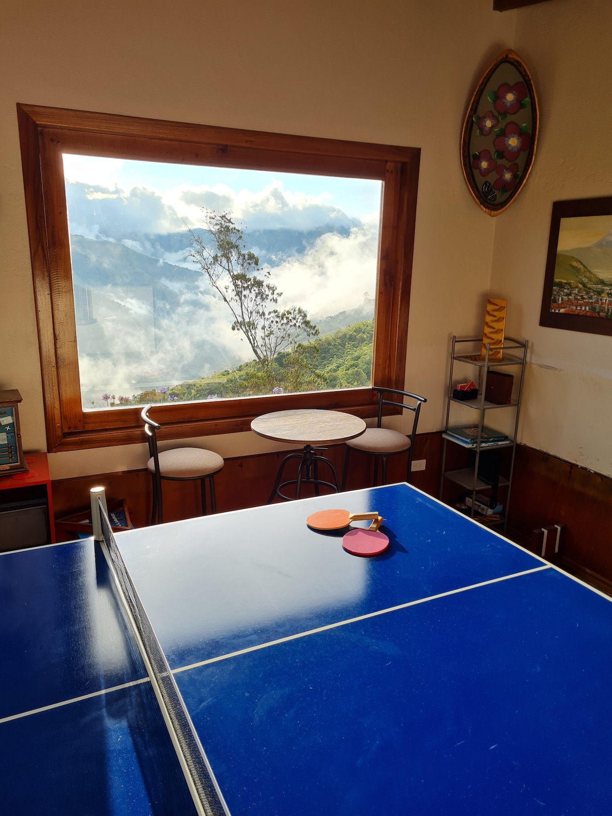 A bright indoor space features a blue ping pong table, positioned beneath a large window that offers a breathtaking view of the surrounding clouds and mountains. Two stools and a small table accompany the game area, enhancing the relaxing atmosphere.