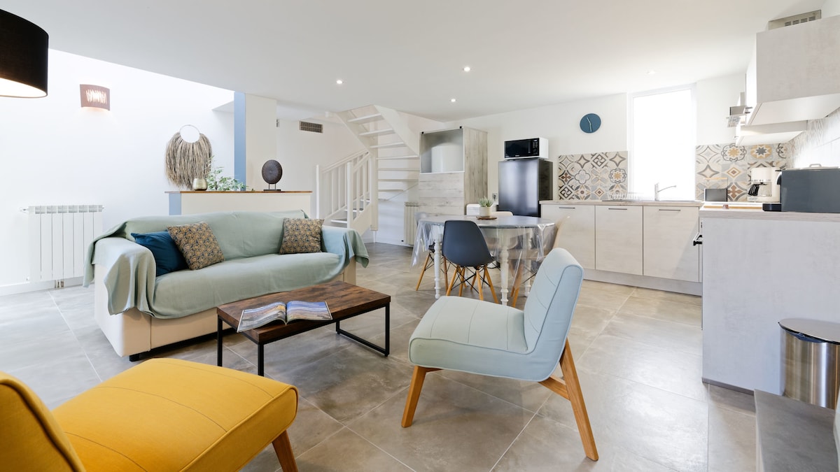 A combined living and kitchen area features a light-colored floor and a staircase leading to the upper level. A large sofa is paired with two accent chairs and a wooden coffee table at the center. The kitchen showcases modern appliances and colorful tile accents.