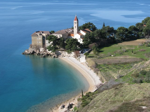 A scenic coastal view displays a small beach bordered by clear blue waters, leading to a charming building with a red-tiled roof and a prominent bell tower. Lush greenery surrounds the structure, enhancing the tranquil setting in the background.