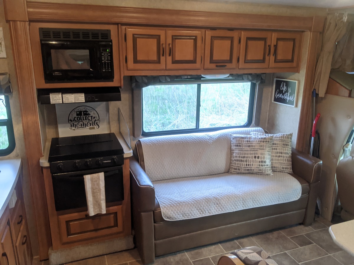 The interior of a camper is displayed, featuring a dual-toned sofa and overhead wooden cabinets. A compact kitchen with a microwave and oven is visible alongside the sofa. A window allows natural light to enter, with a view of greenery outside.