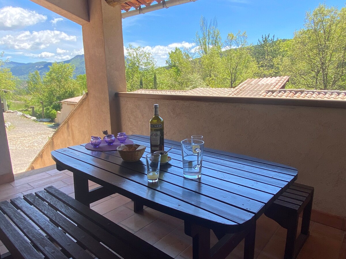 An outdoor terrace is shown with a large wooden table, accompanied by benches. A bottle of olive oil, glasses, and bowls are arranged on the table, with scenic views of trees and distant hills visible in the background under a blue sky.