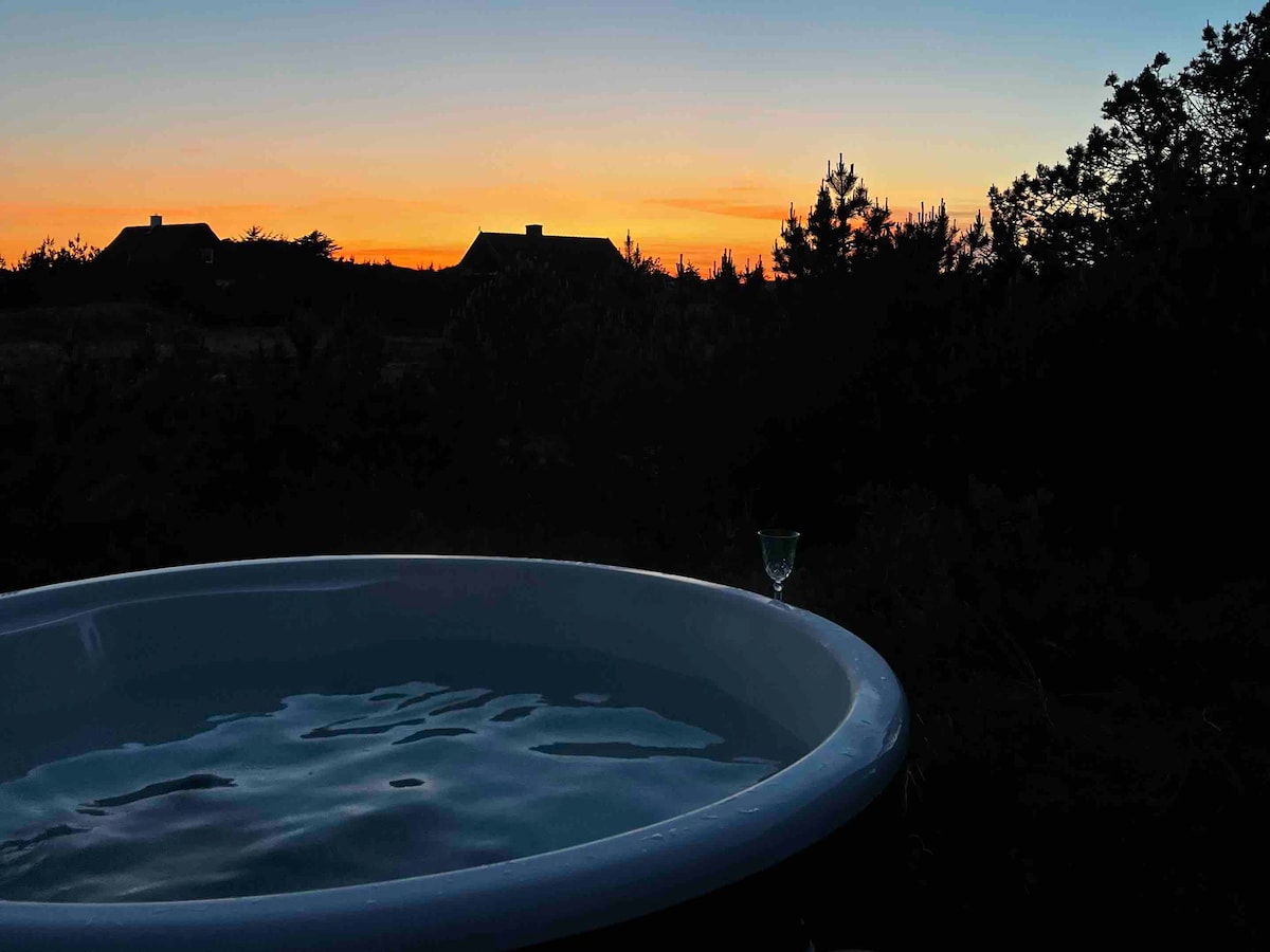 The image captures a warm-lit sunset sky, with hues of orange and purple blending above silhouettes of nearby houses. In the foreground, a round hot tub filled with water reflects the colors, while a glass sits nearby, contributing to a serene atmosphere.