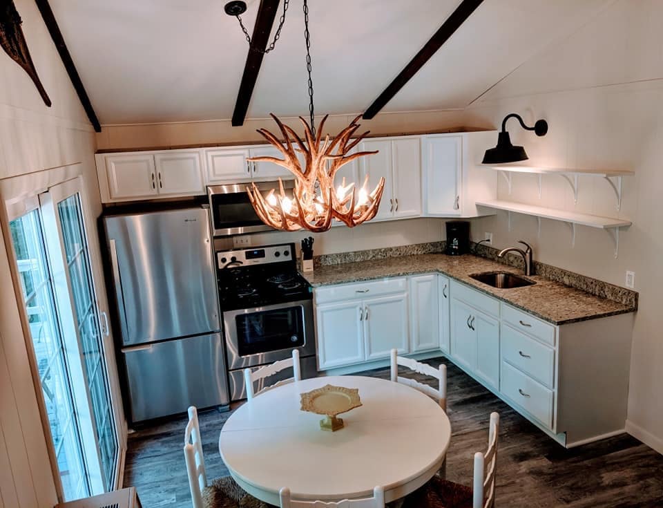 The kitchen features a modern design with stainless steel appliances and granite countertops. A round white dining table is placed at the center, surrounded by four white chairs. An antler chandelier hangs from the ceiling, complementing the exposed wooden beams.