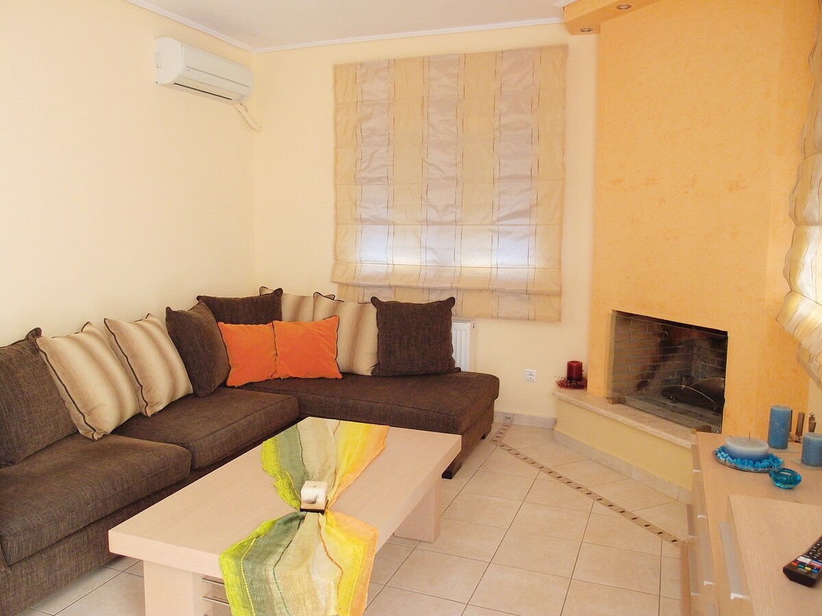 A cozy living area is featured with a spacious sectional sofa adorned with decorative cushions. A coffee table with a colorful runner is positioned in front of the sofa. Soft drapes cover the window, and a fireplace adds a warm touch to the light-filled space.