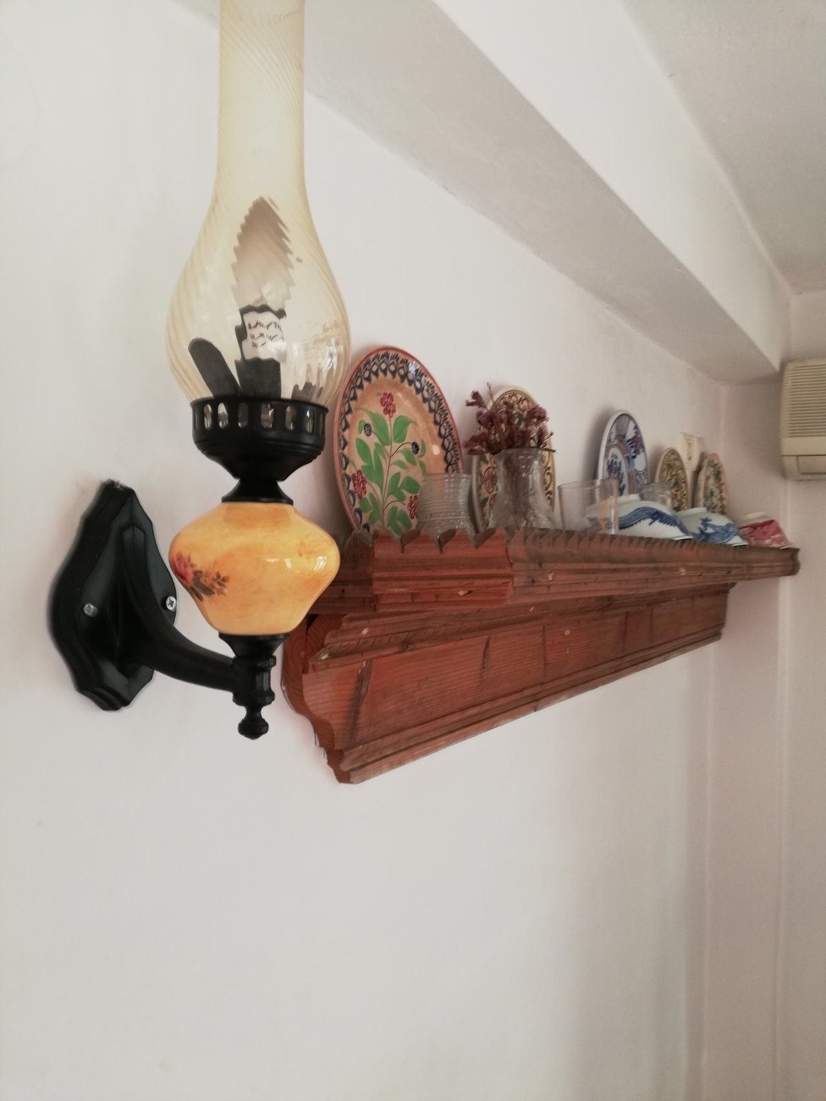A wall-mounted lamp with an amber glass shade is positioned beside a wooden shelf. The shelf holds a variety of decorative plates and glassware, adding a charming touch to the room's decor.