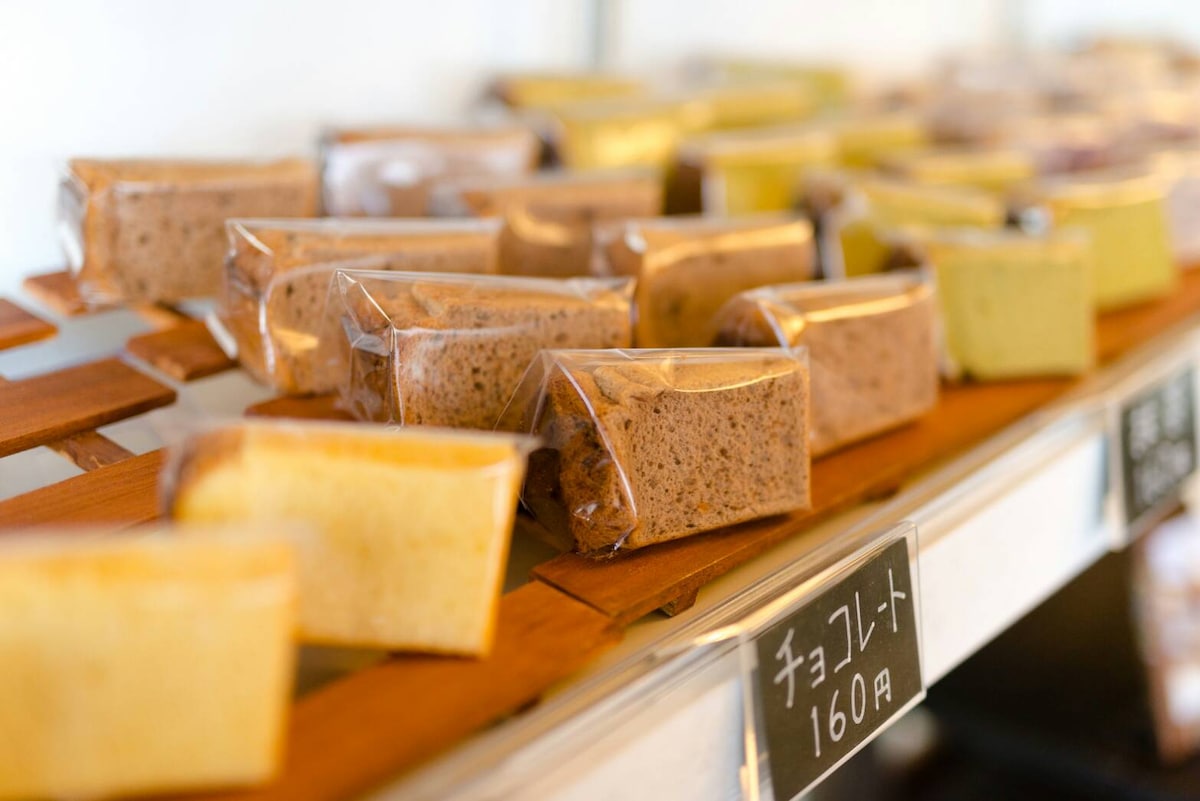 An assortment of neatly packaged chiffon cakes is displayed on wooden shelves. Various flavors are visible, labeled with prices. The cakes feature a soft, moist texture, and the presentation highlights a variety of colors, offering an inviting selection for guests.